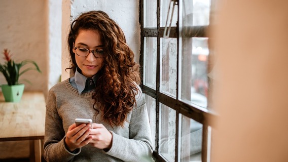 Eine junge Frau mit lockigem Haar schaut intensiv auf ihr Smartphone, das sie mit beiden Händen hält. Im Hintergrund ist ein Wohnraum zu sehen.