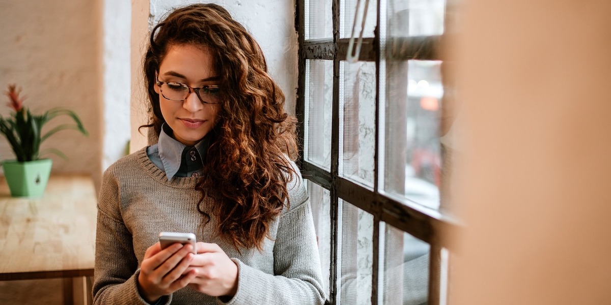 Eine junge Frau mit lockigem Haar schaut intensiv auf ihr Smartphone, das sie mit beiden Händen hält. Im Hintergrund ist ein Wohnraum zu sehen.