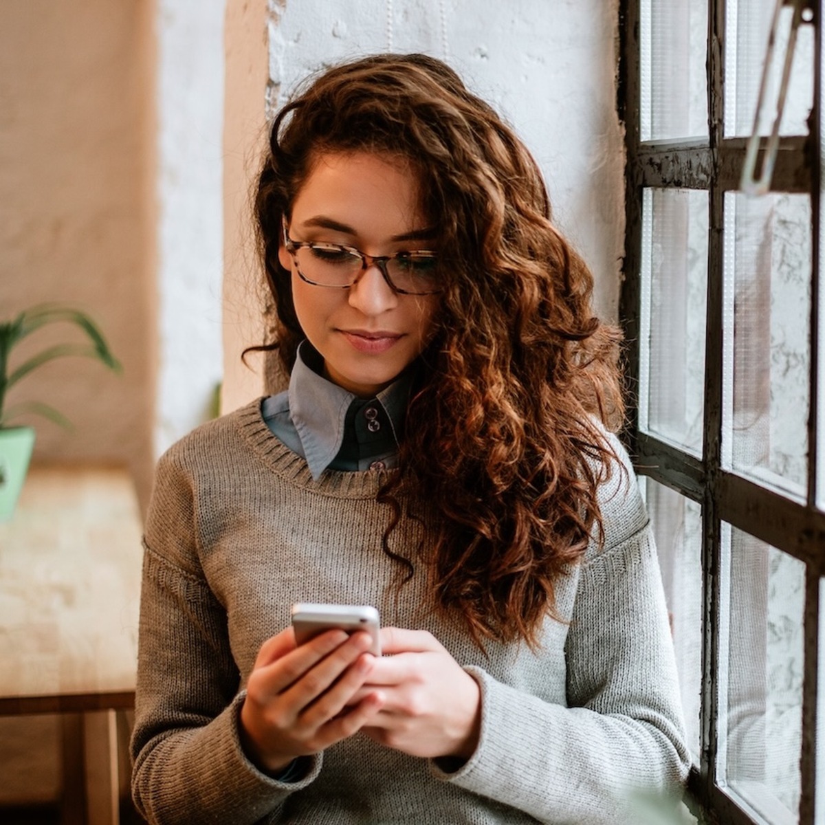 Eine junge Frau mit lockigem Haar schaut intensiv auf ihr Smartphone, das sie mit beiden Händen hält. Im Hintergrund ist ein Wohnraum zu sehen.