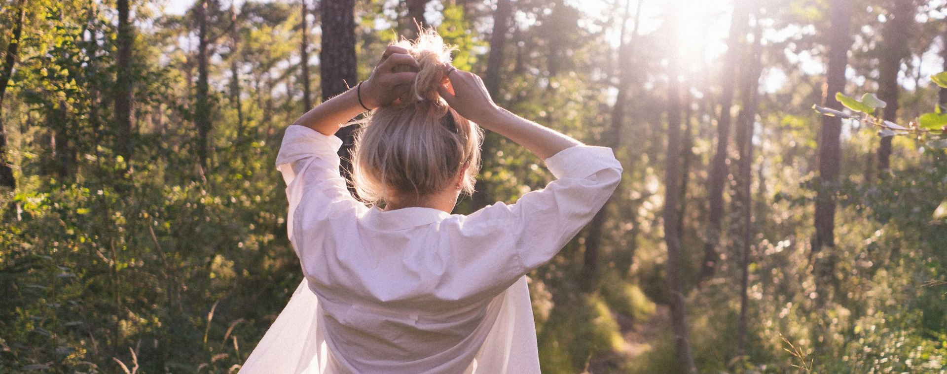 Eine junge Frau beim Spaziergang durch einen lichtdurchfluteten Wald, die Hände in die Haare greifend.