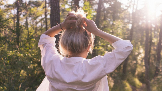 Eine junge Frau beim Spaziergang durch einen lichtdurchfluteten Wald, die Hände in die Haare greifend.