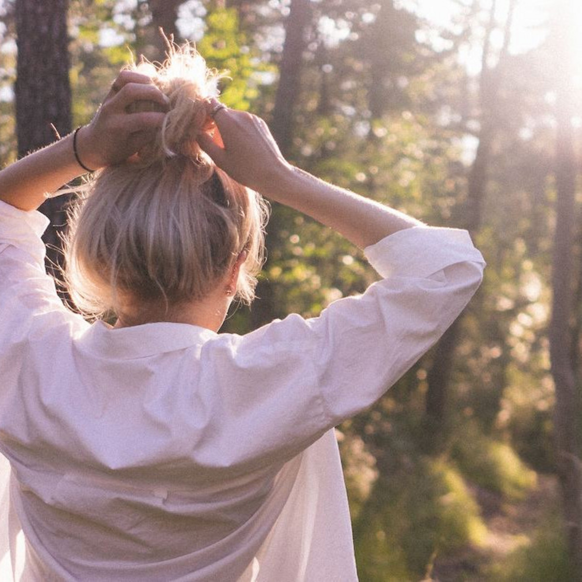 Eine junge Frau beim Spaziergang durch einen lichtdurchfluteten Wald, die Hände in die Haare greifend.