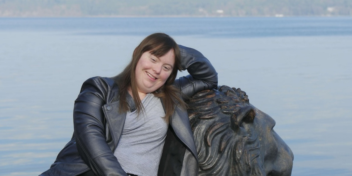 Luisa Wöllisch, eine Frau mit langen braunen Haaren und einem grauen Lederjackett, lehnt sich entspannt an eine Löwenstatue. Im Hintergrund sieht man einen ruhigen See und eine bewaldete Uferlinie. Luisa lächelt und wirkt glücklich.