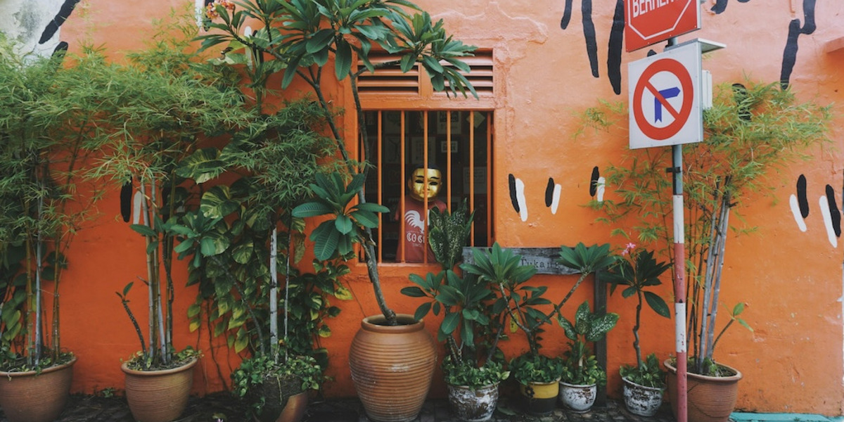 Das Bild zeigt eine farbenfrohe Szene in Melaka, Malaysia. Eine orangefarbene Wand mit künstlerischen schwarzen und weißen Akzenten bildet den Hintergrund. Im Vordergrund stehen mehrere grüne Pflanzen in Töpfen vor der Wand. Ein Fenster mit orangefarbenen Gittern befindet sich in der Mitte des Bildes, dahinter ist eine Figur mit einer goldenen Maske und einem roten Hemd zu sehen. Rechts neben dem Fenster sind zwei Straßenschilder zu erkennen: eines mit dem Wort „Berhenti“ (Malaiisch für „Stopp“), das andere zeigt ein Einfahrverbotsschild. Die gesamte Szene strahlt eine lebendige, urbane Atmosphäre aus.
