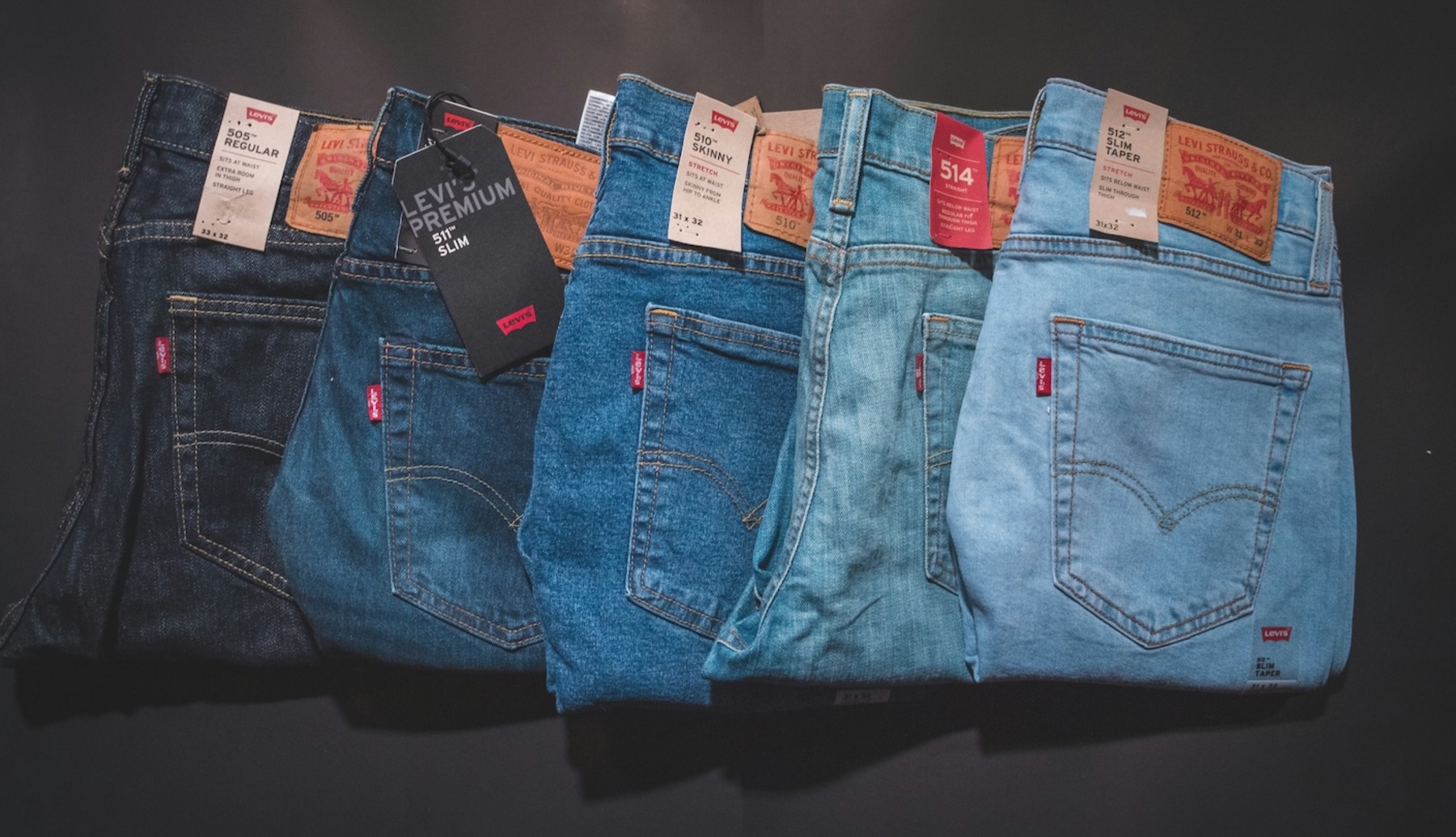 Eine Reihe von gefalteten Jeans in verschiedenen Blautönen liegt nebeneinander, von dunkelblau bis hellblau reichend. Die Hosen zeigen verschiedene Levi's-Lederlabels am Bund und rote Markenlogos an den Gesäßtaschen.