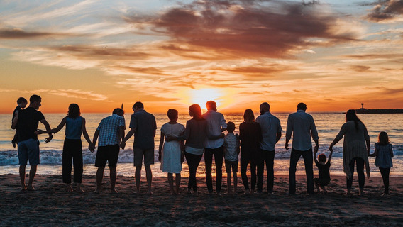 15 Menschen, aller Altersklassen und Geschlechter, stehen am Strand in einer Reihe und halten ihre Hände oder haben sich im Arm. Am Horizont geht die Sonne unter.