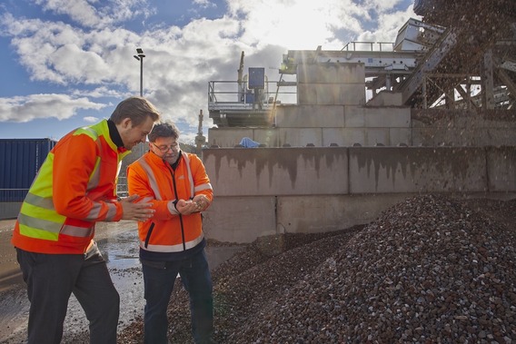 Zwei Personen in orangefarbenen Warnwesten stehen auf einer Baustelle und schauen gemeinsam auf ein Mobilgerät. Im Hintergrund sind Betonwände, Baumaschinen und ein großer Schutthaufen zu sehen.