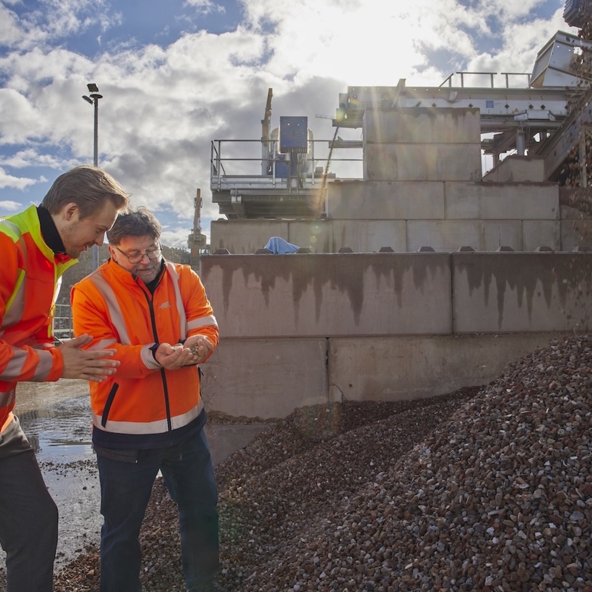 Zwei Personen in orangefarbenen Warnwesten stehen auf einer Baustelle und schauen gemeinsam auf ein Mobilgerät. Im Hintergrund sind Betonwände, Baumaschinen und ein großer Schutthaufen zu sehen.