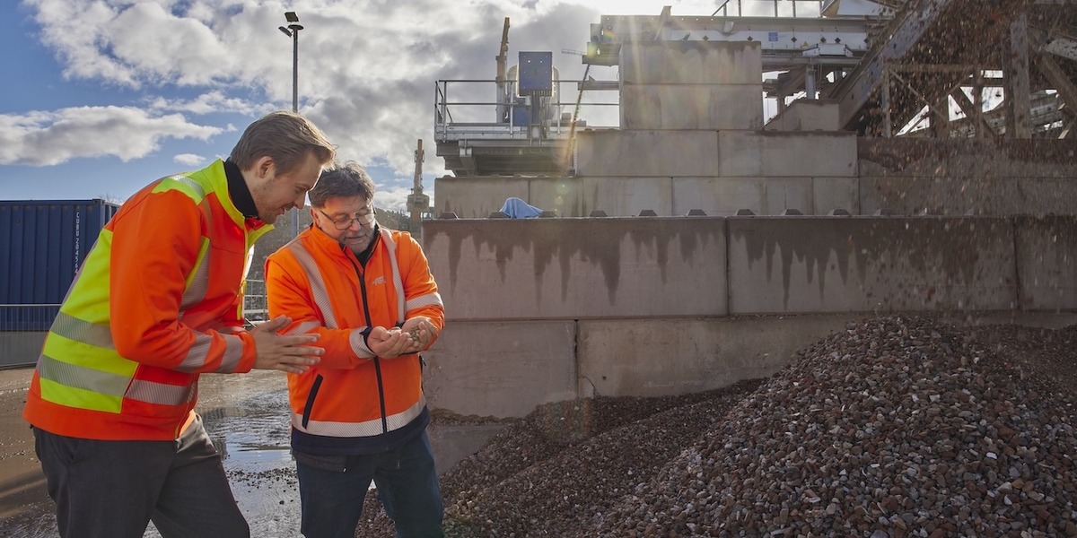 Zwei Personen in orangefarbenen Warnwesten stehen auf einer Baustelle und schauen gemeinsam auf ein Mobilgerät. Im Hintergrund sind Betonwände, Baumaschinen und ein großer Schutthaufen zu sehen.