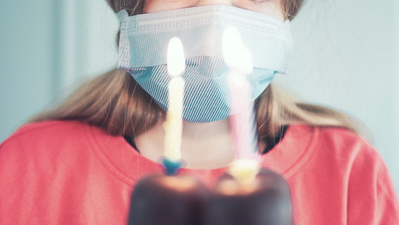 Eine Person mit Maske steht vor einer Torte mit drei Geburtstagskerzen.