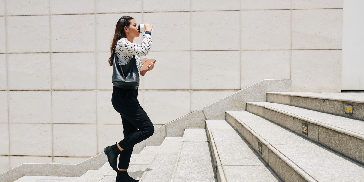 Eine Frau läuft die Treppen hoch und trinkt dabei aus ihrem Kaffee-To-Go-Becher.
