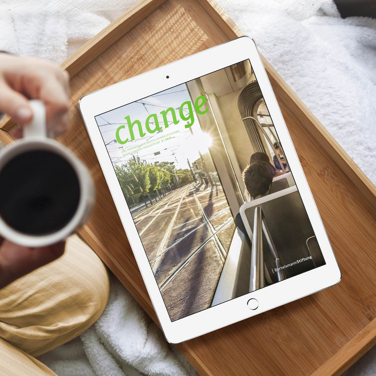 Eine Person sitzt mit einem Kaffee in der Hand vor einem Tablet, auf dem das neue change Magazin der Bertelsmann Stiftung abgebildet ist.
