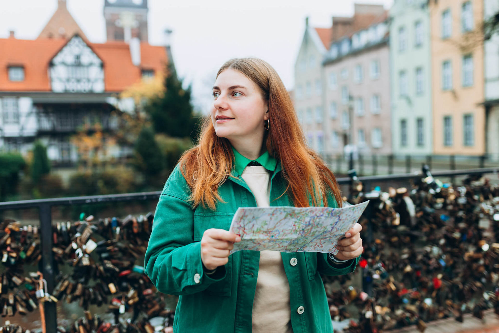 Eine rothaarige, junge Frau in grüner Jacke steht mit einem Stadtplan in der Hand auf einer Brücke in der polnischen Stadt Danzig und schaut sich um.