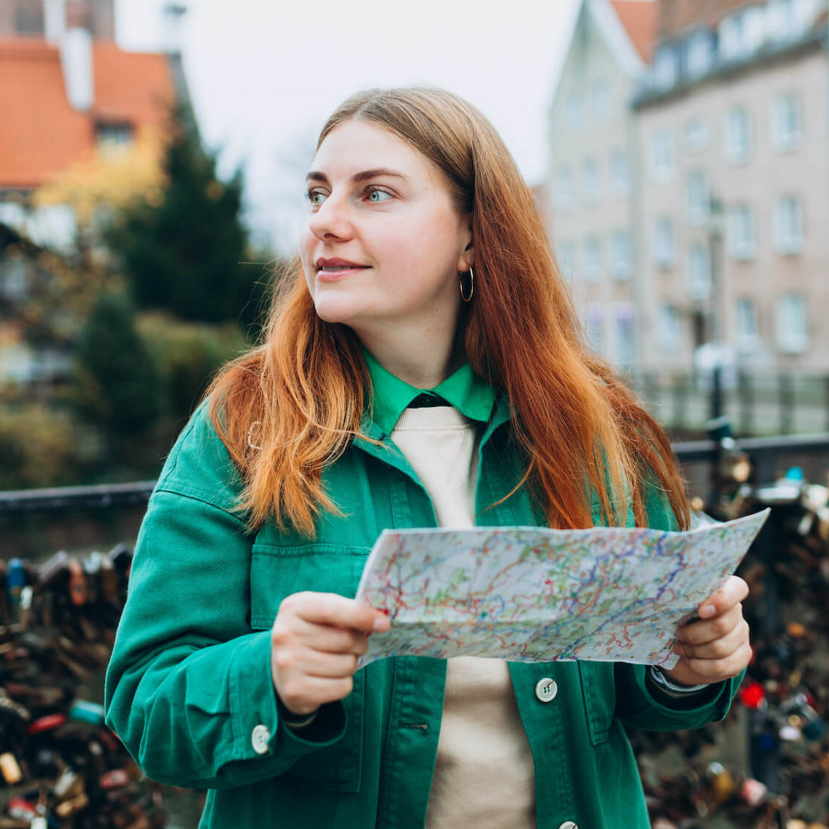 Eine rothaarige, junge Frau in grüner Jacke steht mit einem Stadtplan in der Hand auf einer Brücke in der polnischen Stadt Danzig und schaut sich um.