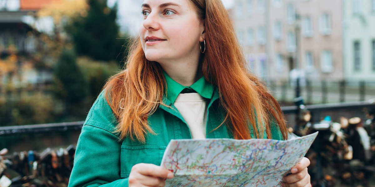 Eine rothaarige, junge Frau in grüner Jacke steht mit einem Stadtplan in der Hand auf einer Brücke in der polnischen Stadt Danzig und schaut sich um.