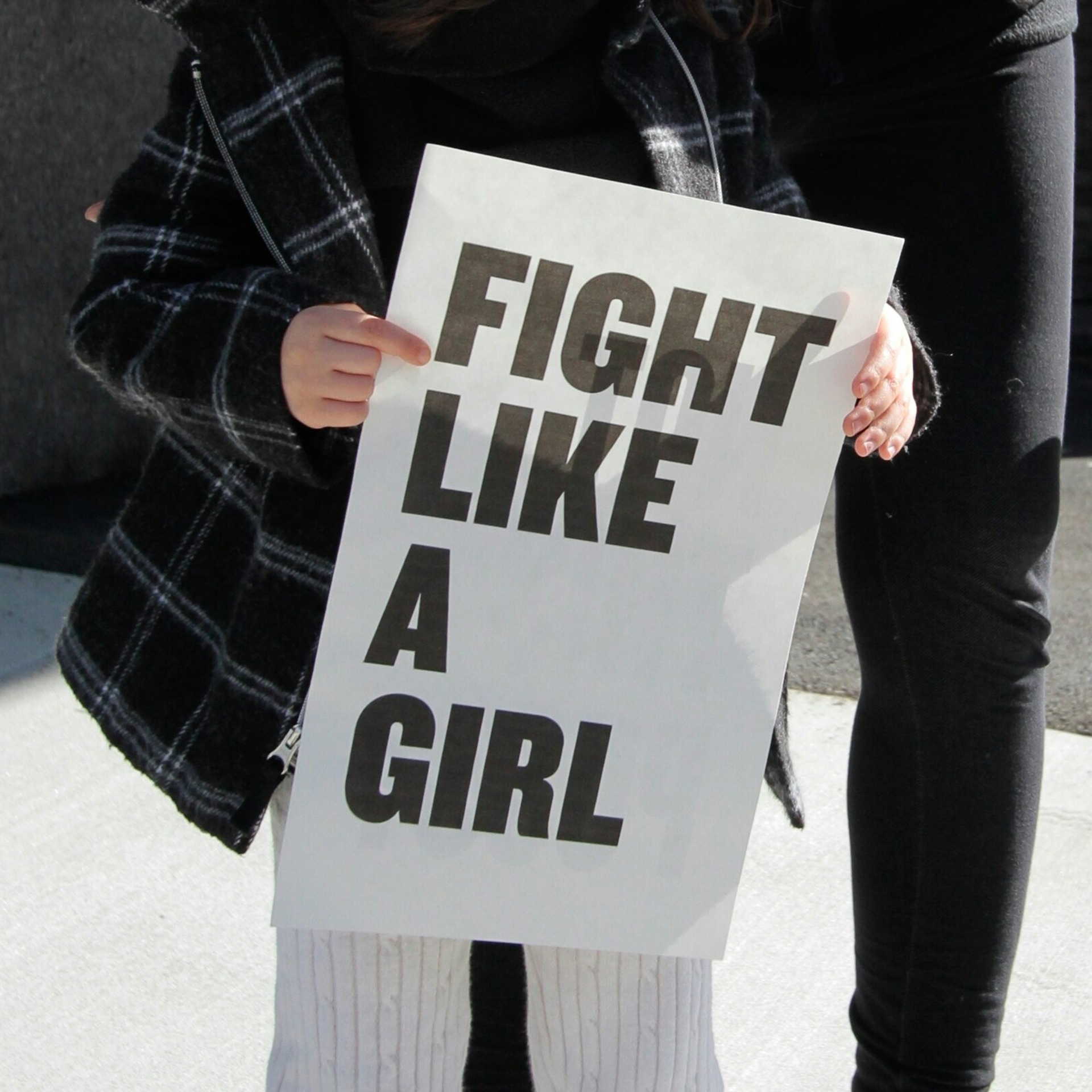 Eine Person hält bei einer Demonstration ein weißes Schild mit der Aufschrift „Fight Like A Girl" in den Händen, im Hintergrund steht eine weitere Person auf dem Gehweg.