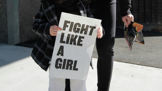 Eine Person hält bei einer Demonstration ein weißes Schild mit der Aufschrift „Fight Like A Girl" in den Händen, im Hintergrund steht eine weitere Person auf dem Gehweg.