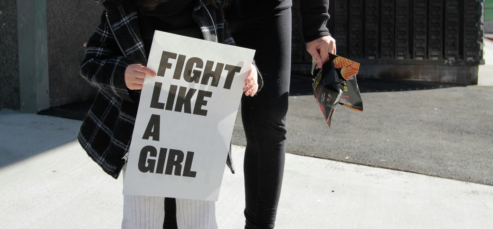 Eine Person hält bei einer Demonstration ein weißes Schild mit der Aufschrift „Fight Like A Girl" in den Händen, im Hintergrund steht eine weitere Person auf dem Gehweg.