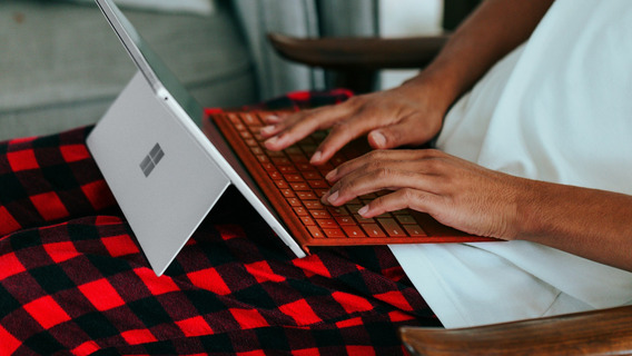 Nahaufnahme von Händen, die auf einer orangefarbenen Tastatur eines Microsoft Surface Laptops tippen. Der Laptop steht auf einer rot-schwarz karierten Decke, die über einem Holzstuhl liegt. Im Hintergrund ist der weiße Bildschirm des aufgeklappten Laptops zu sehen.