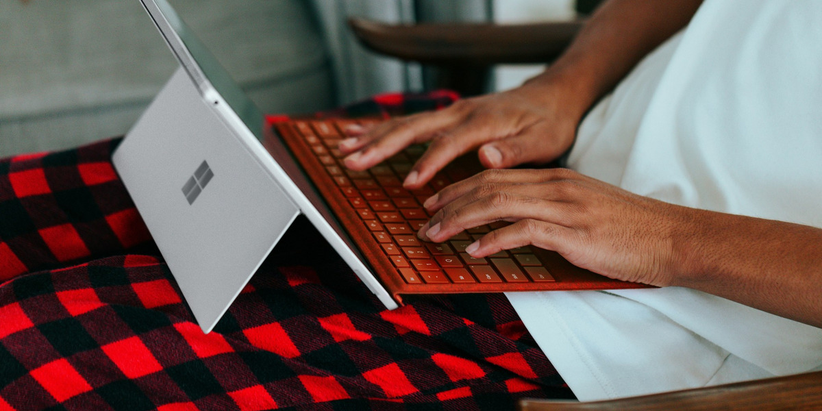 Nahaufnahme von Händen, die auf einer orangefarbenen Tastatur eines Microsoft Surface Laptops tippen. Der Laptop steht auf einer rot-schwarz karierten Decke, die über einem Holzstuhl liegt. Im Hintergrund ist der weiße Bildschirm des aufgeklappten Laptops zu sehen.