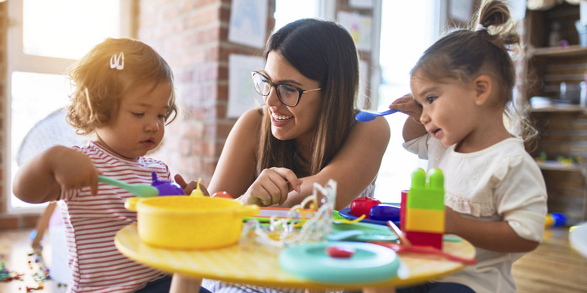 Eine junge Frau spielt lachend mit zwei Kindern in einer Kindertagesstätte