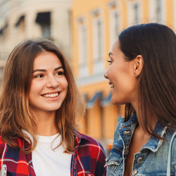 Zwei junge Frauen stehen auf einem Platz und lächeln sich an.