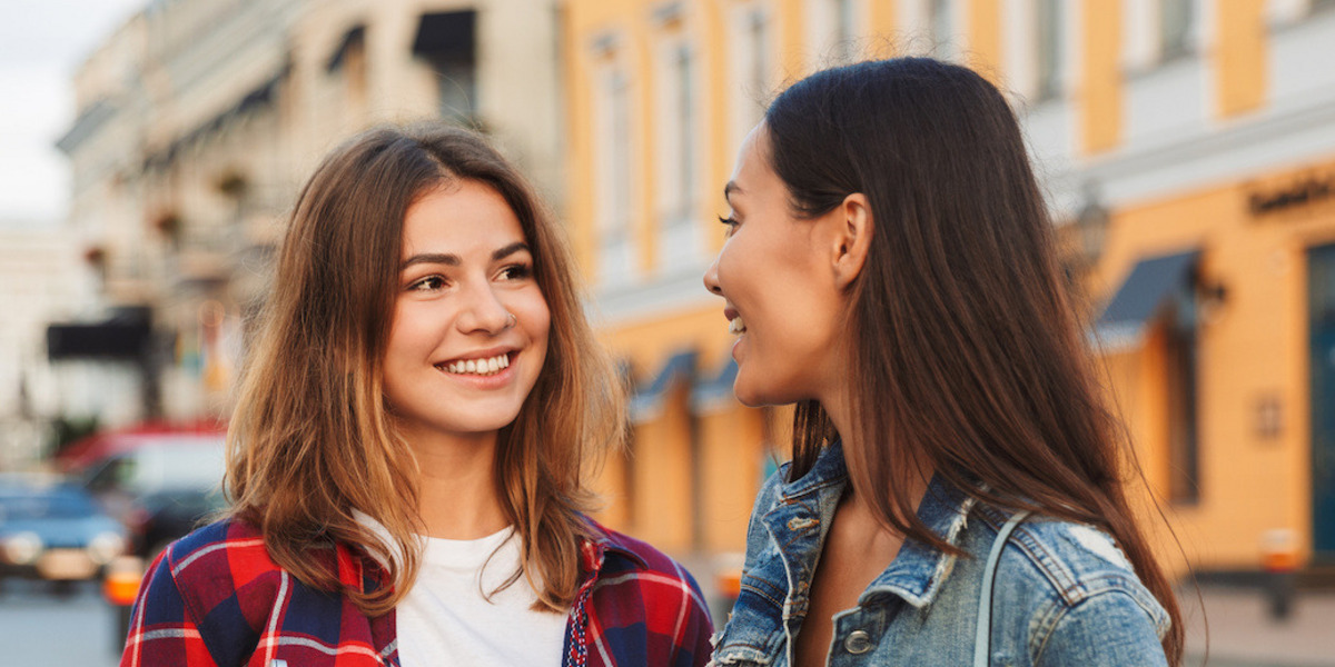 Zwei junge Frauen stehen auf einem Platz und lächeln sich an.