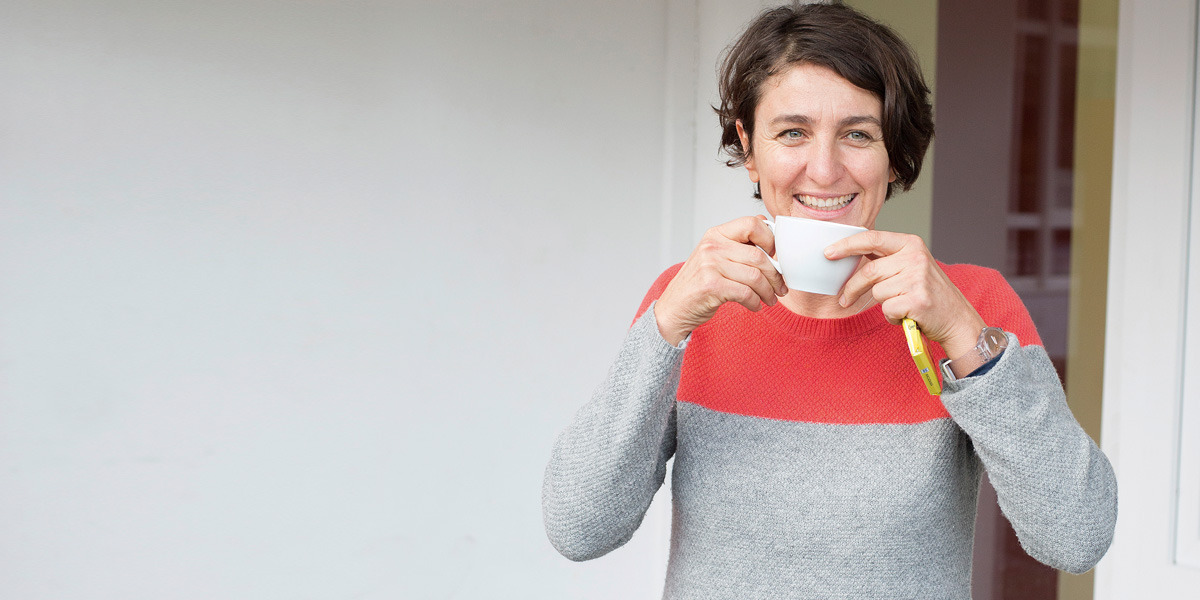 Christiane Falge trinkt aus einer Kaffeetasse und lacht in die Kamera.