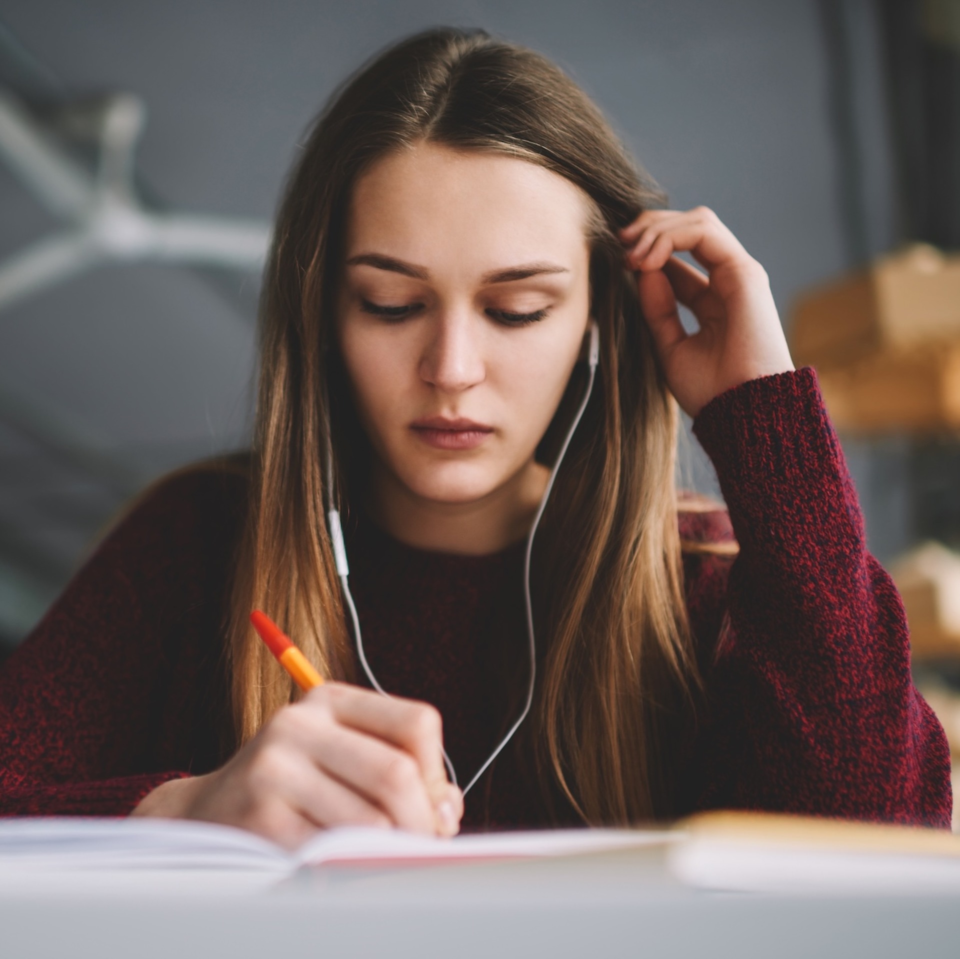 Eine junge Frau sitzt an einem Tisch, hört über Kopfhörer und schreibt konzentriert mit einem Stift in ein aufgeschlagenes Notizbuch.