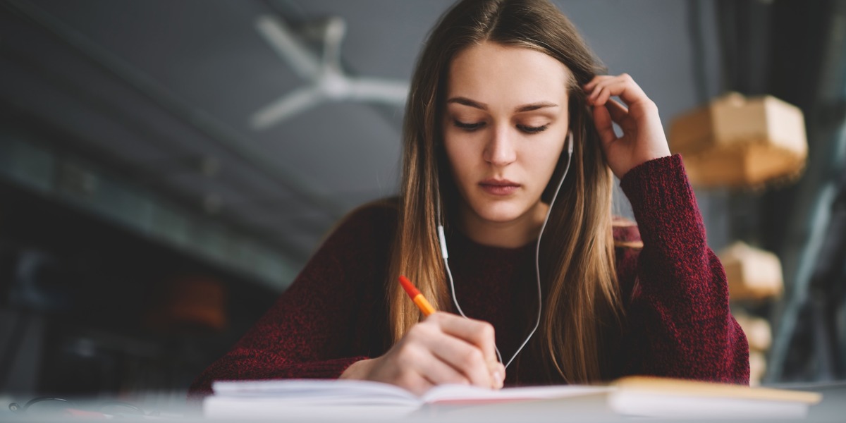 Eine junge Frau sitzt an einem Tisch, hört über Kopfhörer und schreibt konzentriert mit einem Stift in ein aufgeschlagenes Notizbuch.