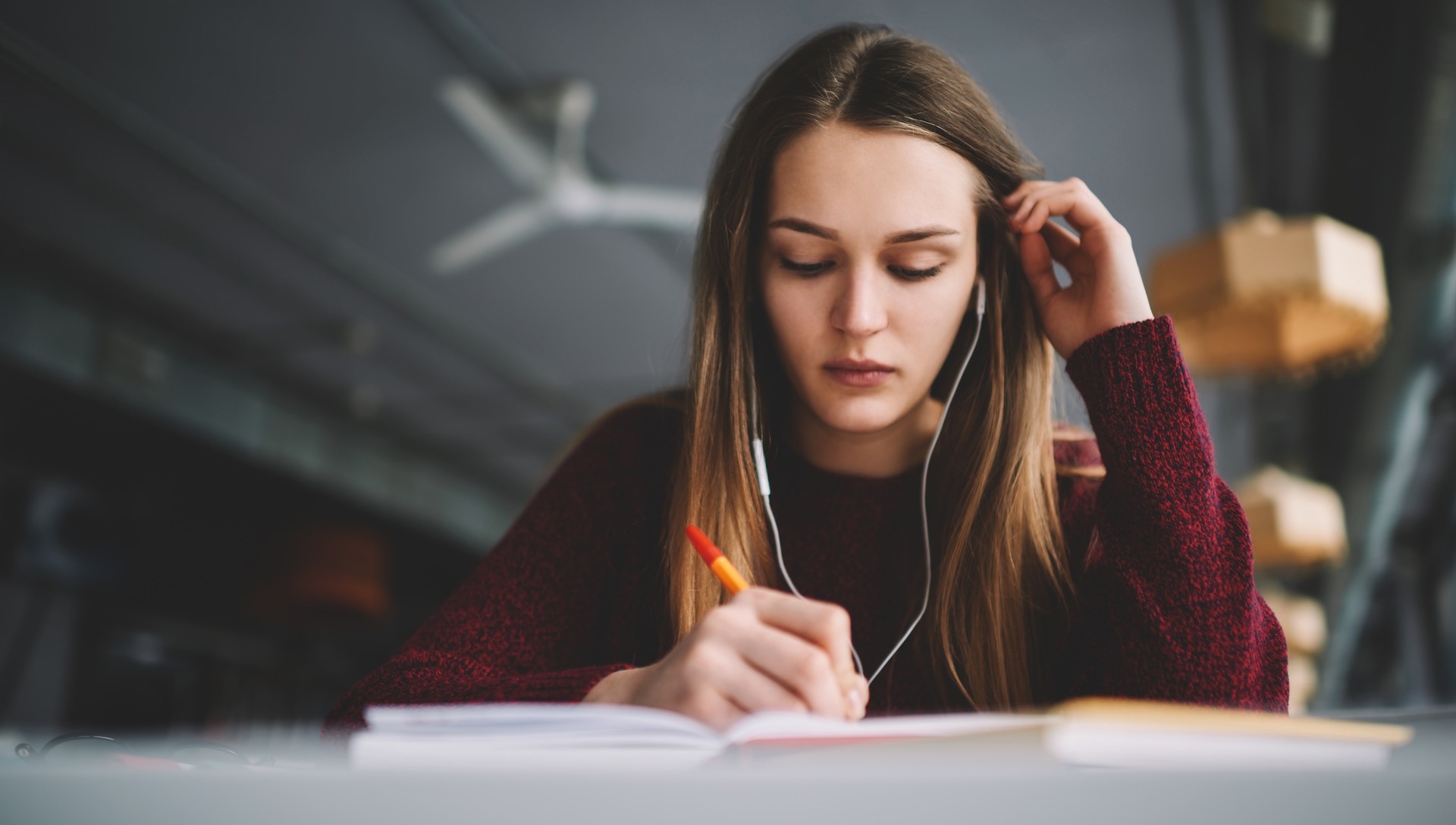 Eine junge Frau sitzt an einem Tisch, hört über Kopfhörer und schreibt konzentriert mit einem Stift in ein aufgeschlagenes Notizbuch.