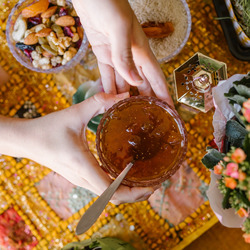 Auf einem Tisch mit einer bunten Tischdecke steht eine Blume, sowie Gefäße mit Lebensmitteln, darunter Nüsse und Reis. Zwei Hände halten eine Schale, in die ein Gelee gefüllt ist, darin liegt ein Löffel.