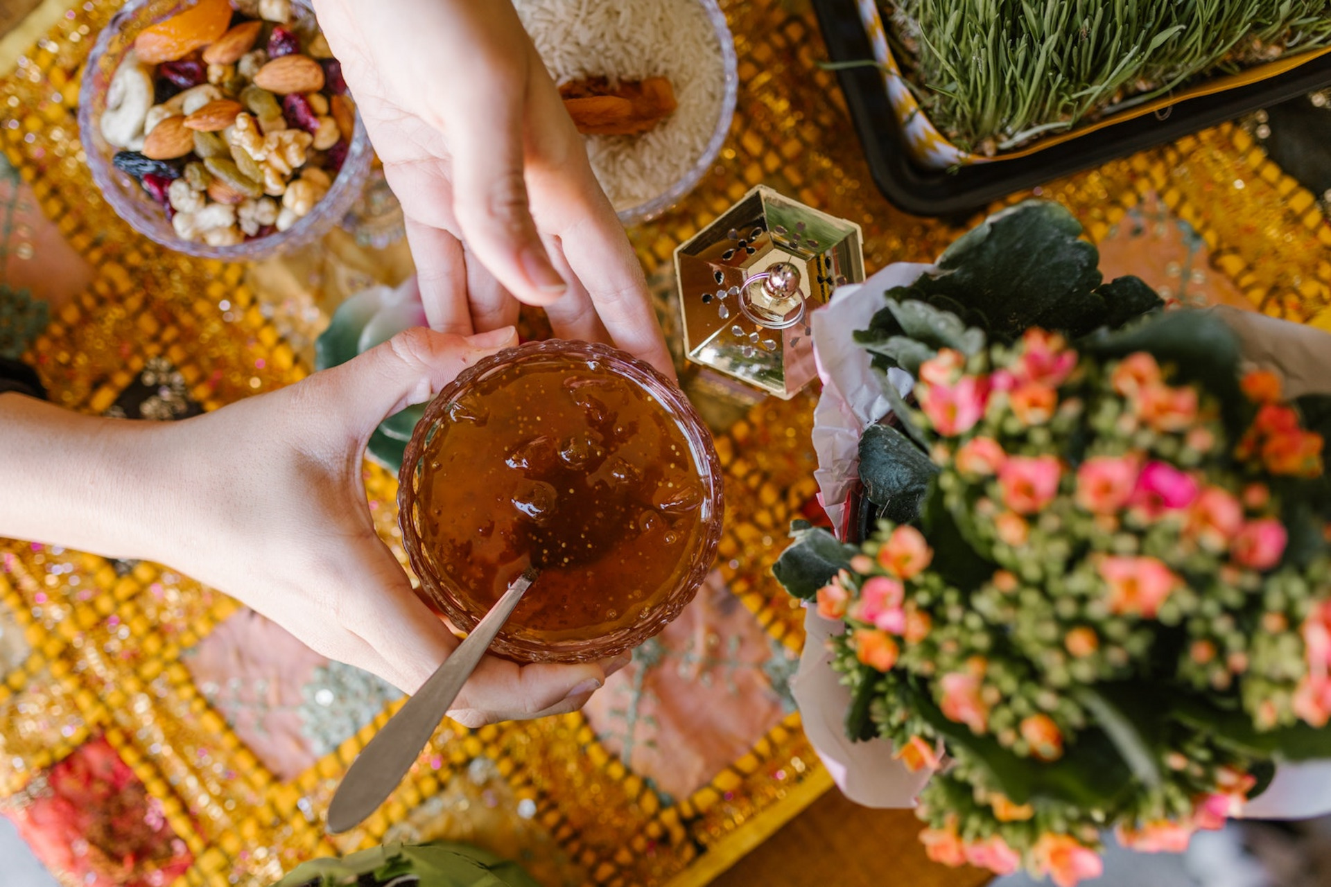 Auf einem Tisch mit einer bunten Tischdecke steht eine Blume, sowie Gefäße mit Lebensmitteln, darunter Nüsse und Reis. Zwei Hände halten eine Schale, in die ein Gelee gefüllt ist, darin liegt ein Löffel.