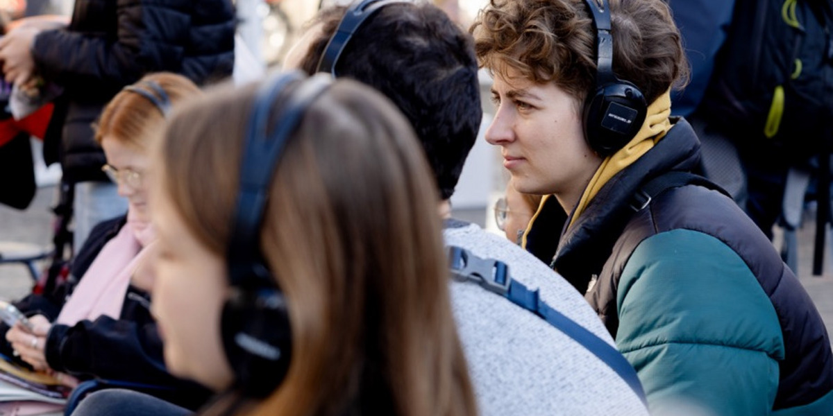 Eine belebte Außenszene mit sitzenden und stehenden Menschen. Im Vordergrund sitzen mehrere junge Leute mit Kopfhörern. Eine Person mit lockigem Haar und grün-schwarzer Jacke schaut konzentriert und trägt gelbe Kopfhörer. Andere um sie herum tragen ebenfalls Kopfhörer, was auf eine Audio-Tour oder -Veranstaltung hindeutet. Im Hintergrund sieht man einen geschäftigen Bereich mit weiteren gehenden und stehenden Menschen.