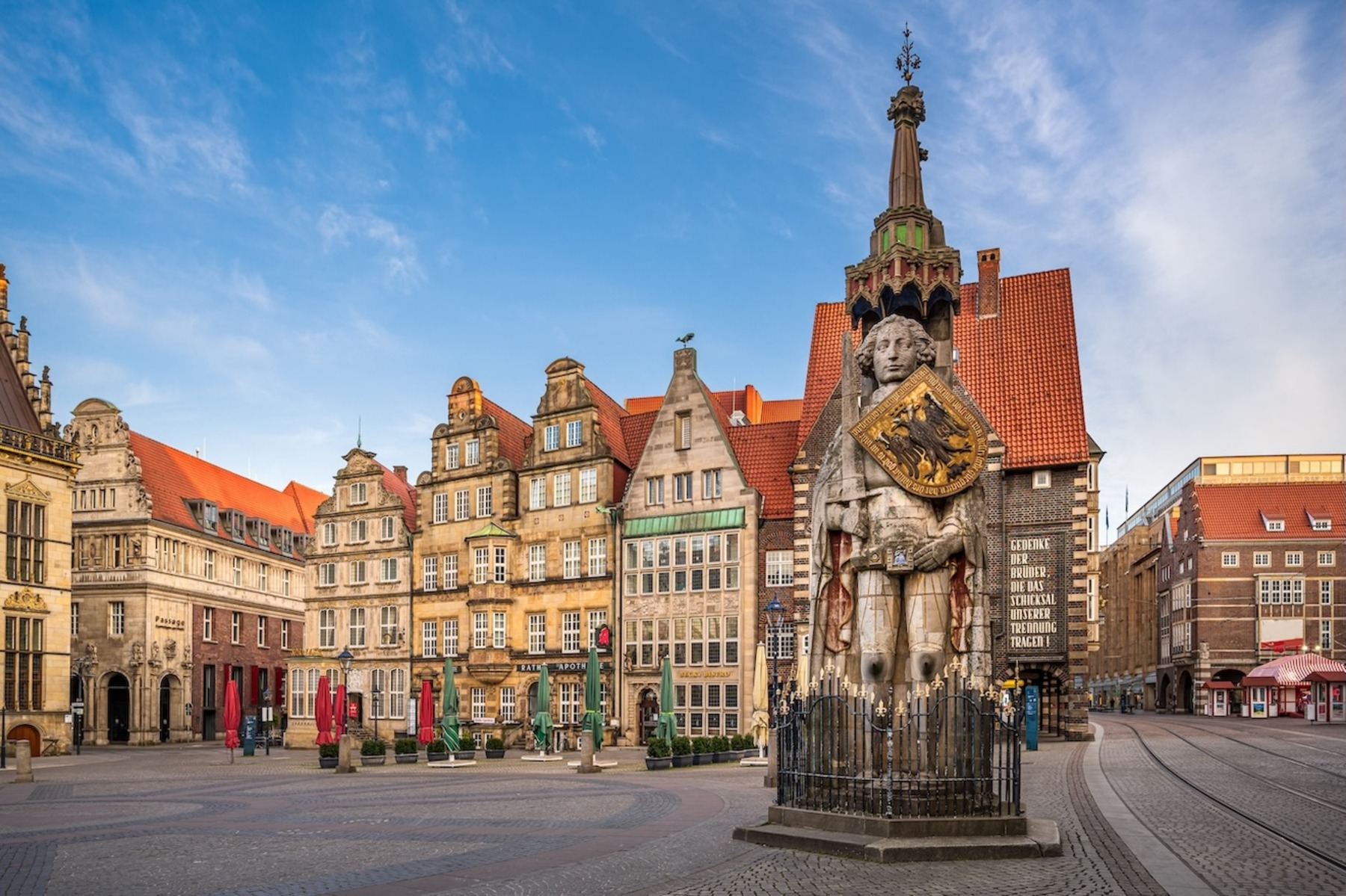 Die Rolandstatue auf dem historischen Marktplatz in Bremen, umgeben von traditionellen norddeutschen Gebäuden mit roten Ziegeldächern und Giebeln unter einem klaren blauen Himmel.