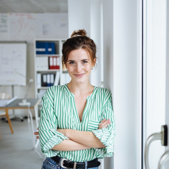 Eine junge Frau lehnt in einem Büro lächelnd an der Wand