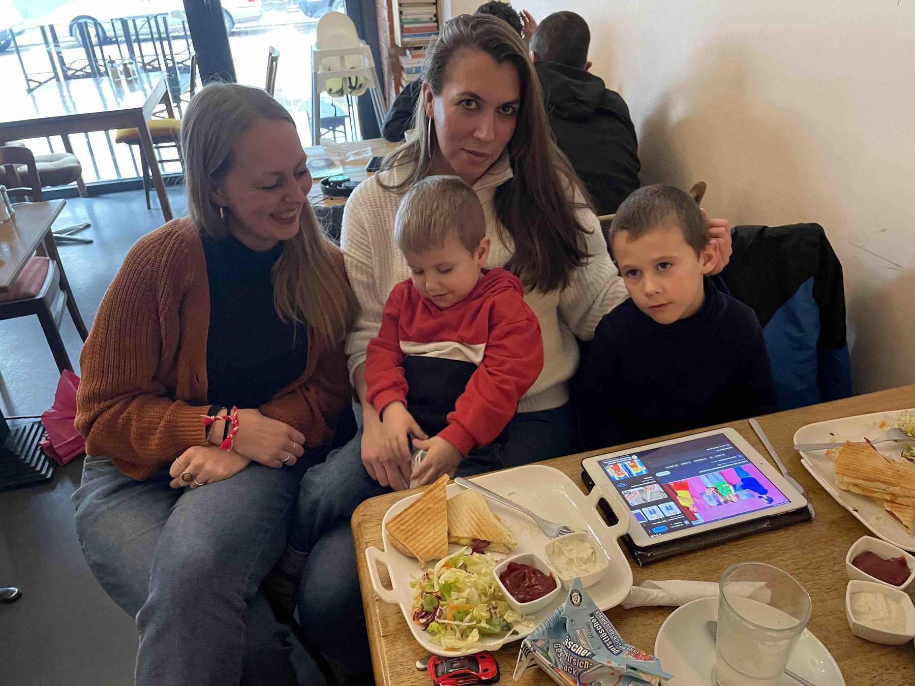 Maren, Hanna und ihre beiden Söhne sitzen an einem Tisch, auf dem Teller mit Essen und ein Glas steht. Daneben liegt ein Tablet.