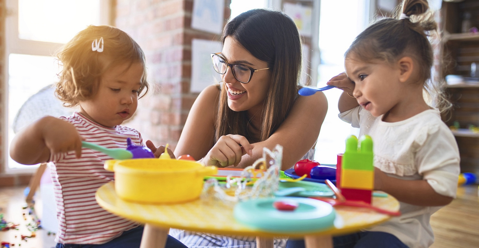 Eine junge Frau spielt lachend mit zwei Kindern in einer Kindertagesstätte