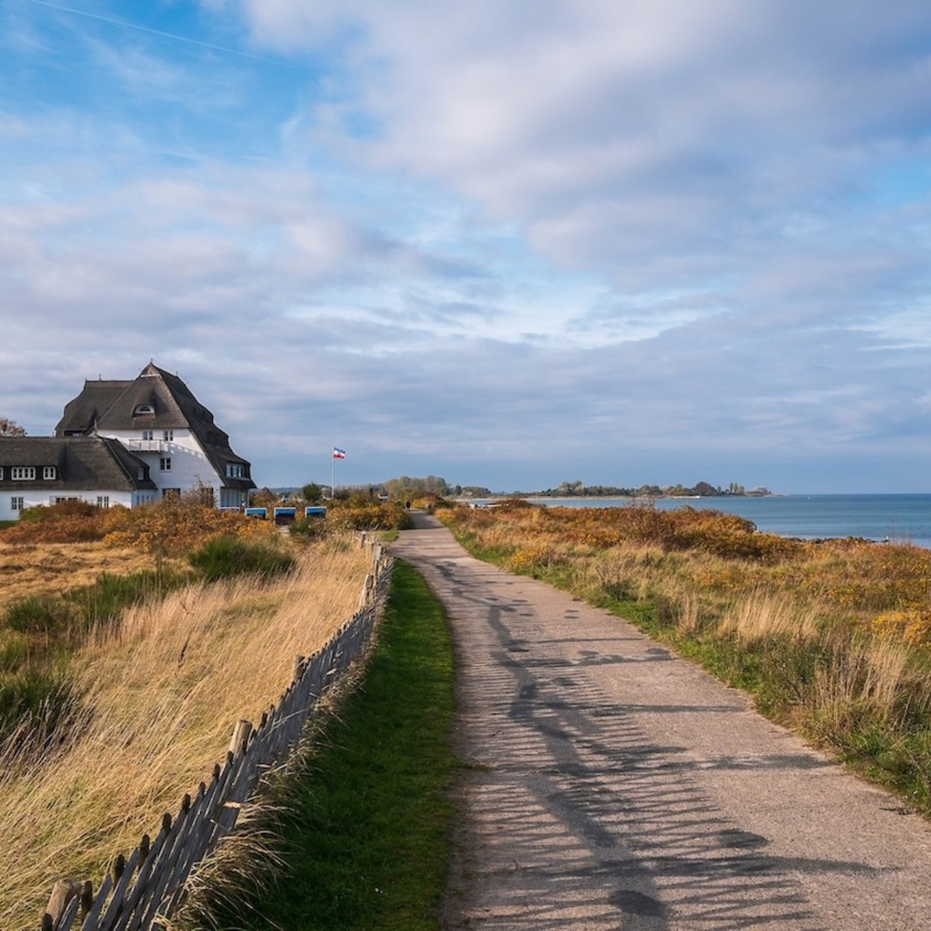 Ein sandiger Weg führt entlang einer Küstenlandschaft mit trockenem Gras und Büschen, rechts das Meer und links ein weißes Haus mit Reetdach unter einem bewölkten Himmel.
