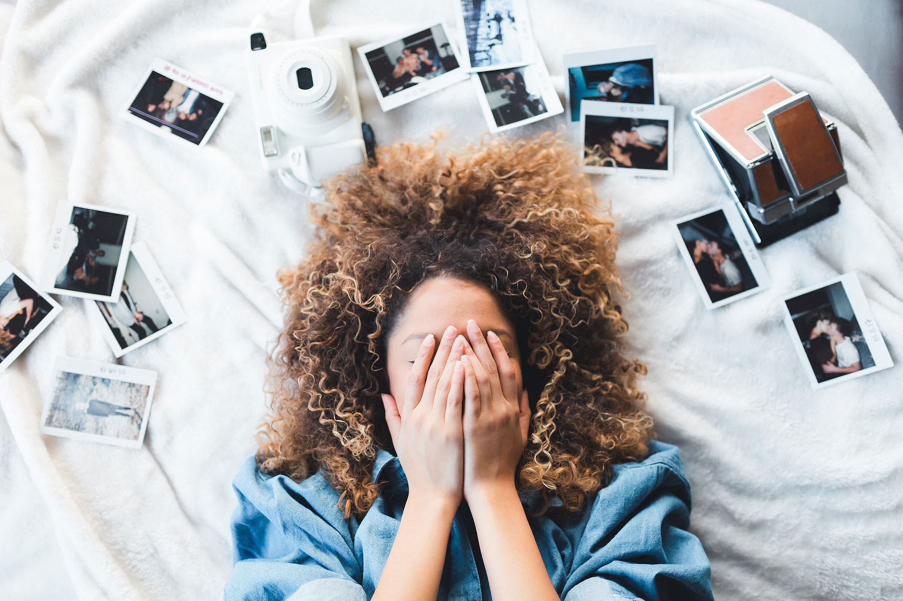 Eine Frau mit lockigen Haaren liegt auf einem Bett, umgeben von Polaroidfotos. Sie verdeckt sich das Gesicht-