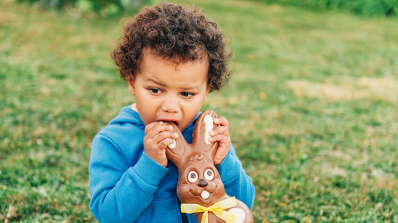 Ein kleiner Junge sitzt im Freien auf dem Rasen und knabbert einem Osterhasen aus Schokolade am Ohr