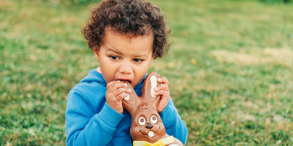 Ein kleiner Junge sitzt im Freien auf dem Rasen und knabbert einem Osterhasen aus Schokolade am Ohr