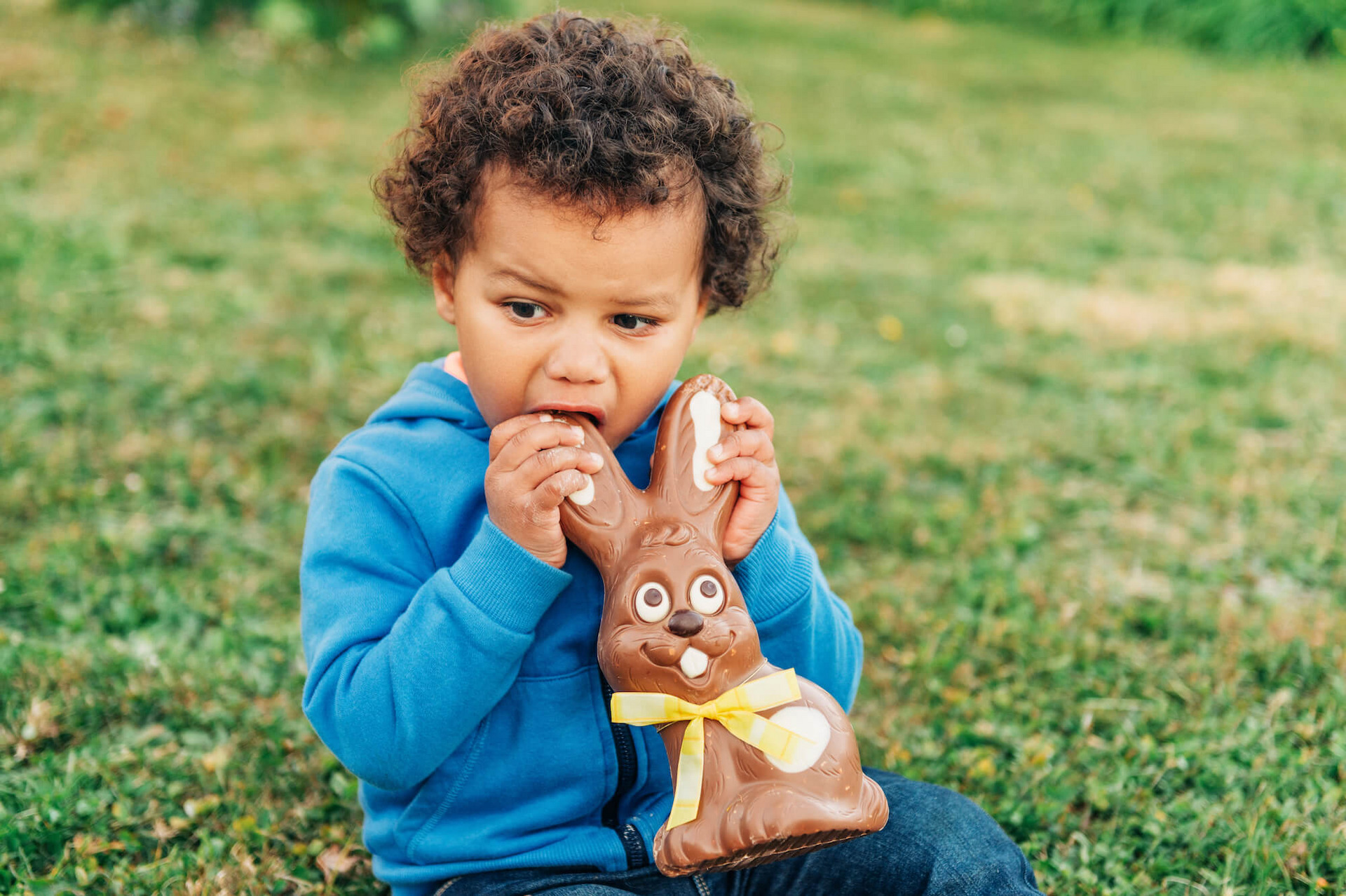 Ein kleiner Junge sitzt im Freien auf dem Rasen und knabbert einem Osterhasen aus Schokolade am Ohr