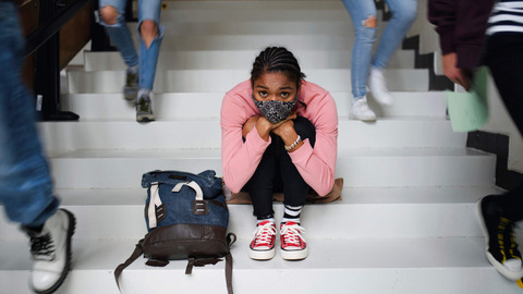 Eine junge Frau mit Alltagsmaske sitzt auf den Treppenstufen in der Schule und schaut besorgt. Neben ihr laufen Schulkamerad:innen die Treppe hinab.