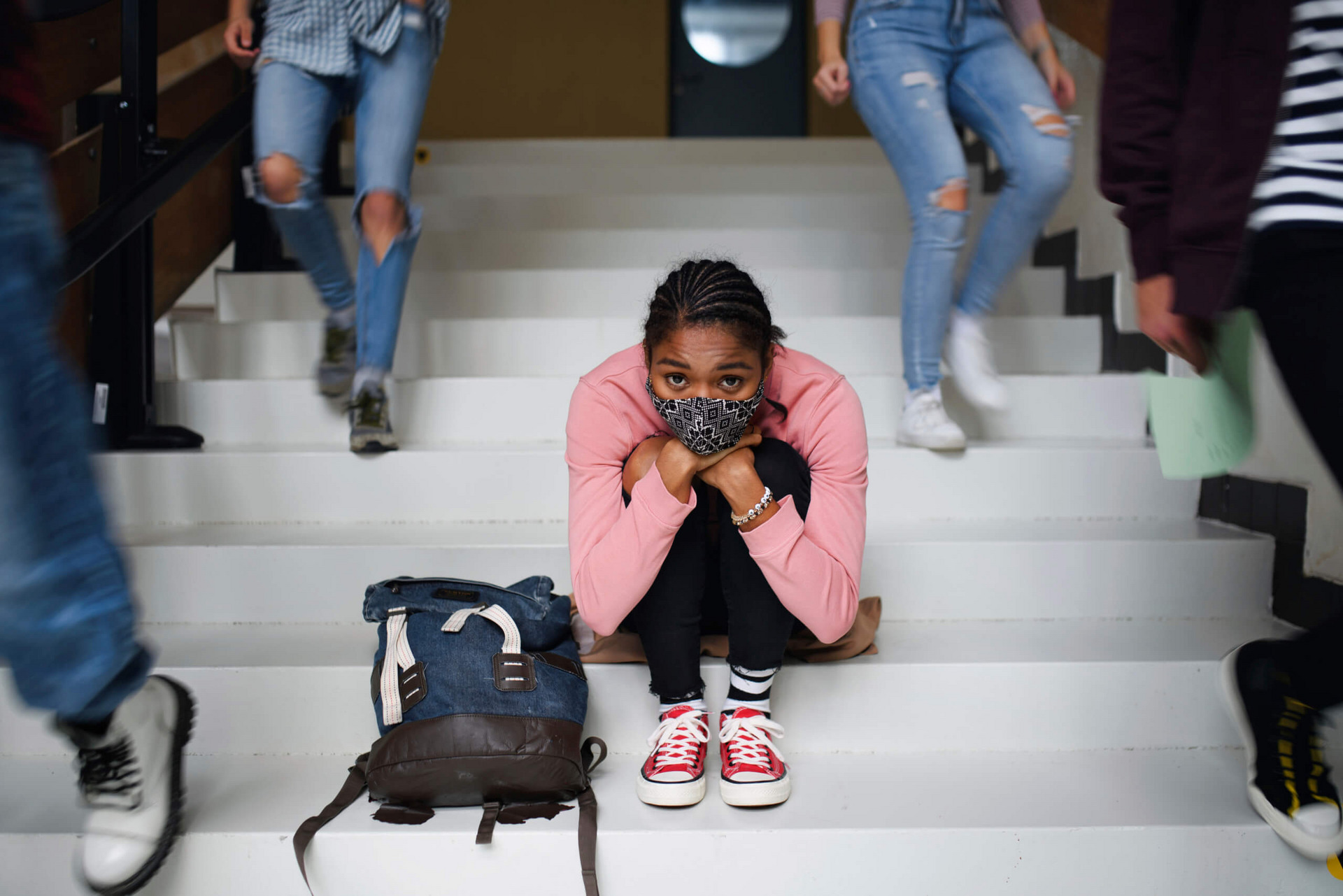 Eine junge Frau mit Alltagsmaske sitzt auf den Treppenstufen in der Schule und schaut besorgt. Neben ihr laufen Schulkamerad:innen die Treppe hinab.