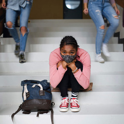 Eine junge Frau mit Alltagsmaske sitzt auf den Treppenstufen in der Schule und schaut besorgt. Neben ihr laufen Schulkamerad:innen die Treppe hinab.