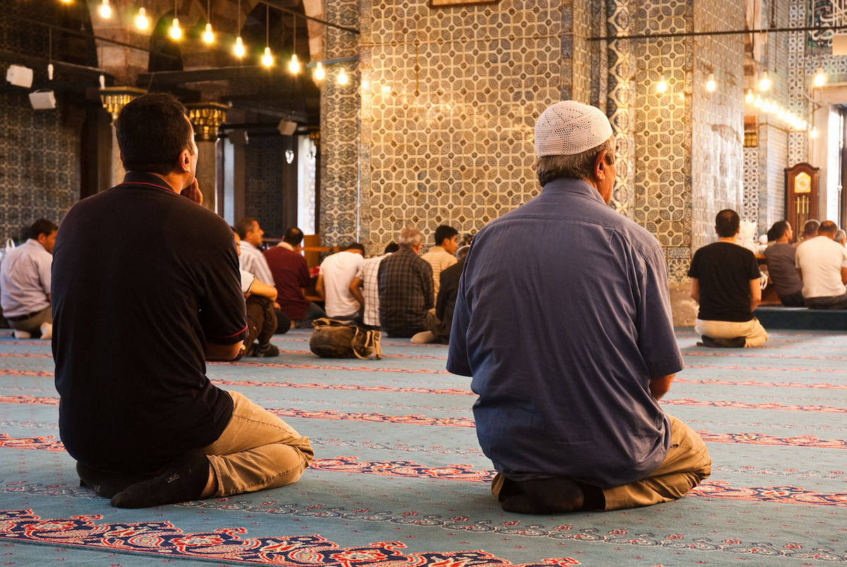 Mit dem Rücken zu uns gewandte Personen beten auf den Knien in einer hell erleuchteten Moschee.
