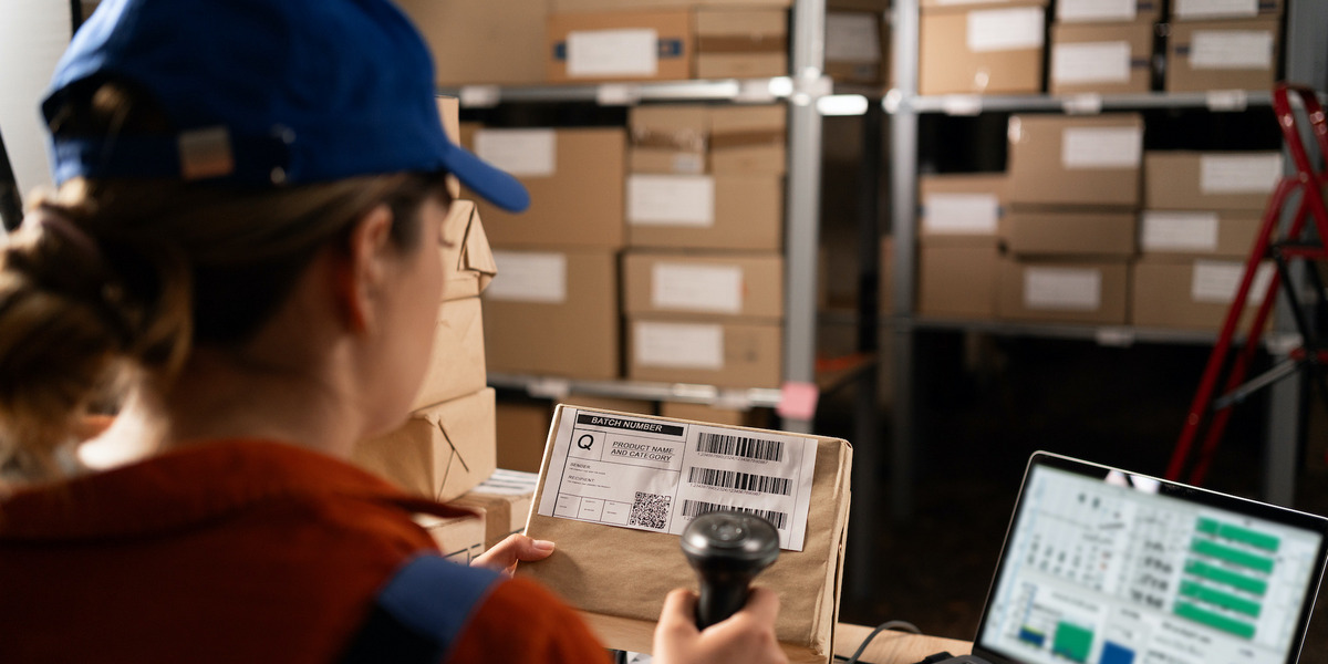 Eine Frau in einem Lager scannt Codes von einem Paketaufkleber, im Hintergrund viele Kisten in großen Regalen, neben ihr ein Laptop mit technischen Daten.