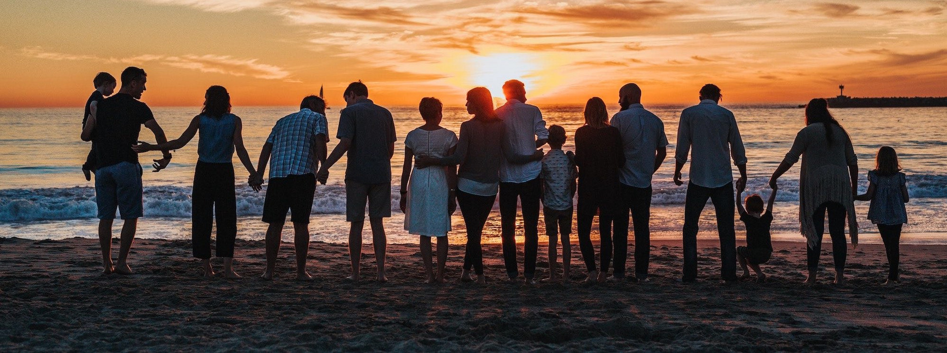 15 Menschen, aller Altersklassen und Geschlechter, stehen am Strand in einer Reihe und halten ihre Hände oder haben sich im Arm. Am Horizont geht die Sonne unter.