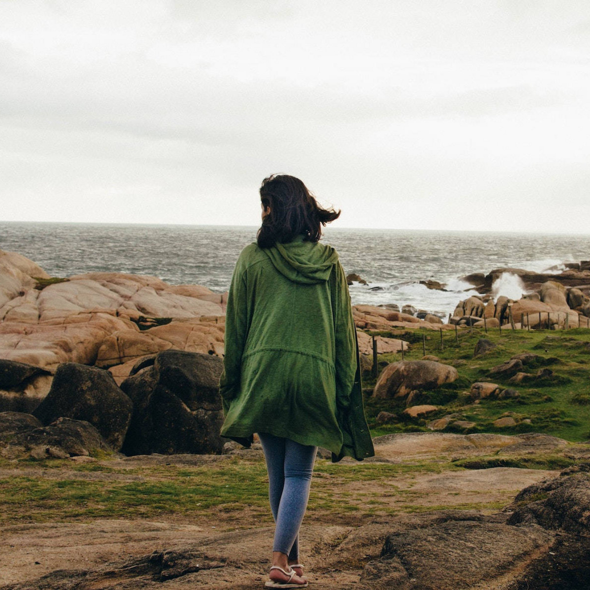 Eine Person in einem grünen Mantel läuft, uns den Rücken zugekehrt, den felsigen Strand von Cabo Polonio in Uruguay entlang, auf den tosende Wellen treffen.